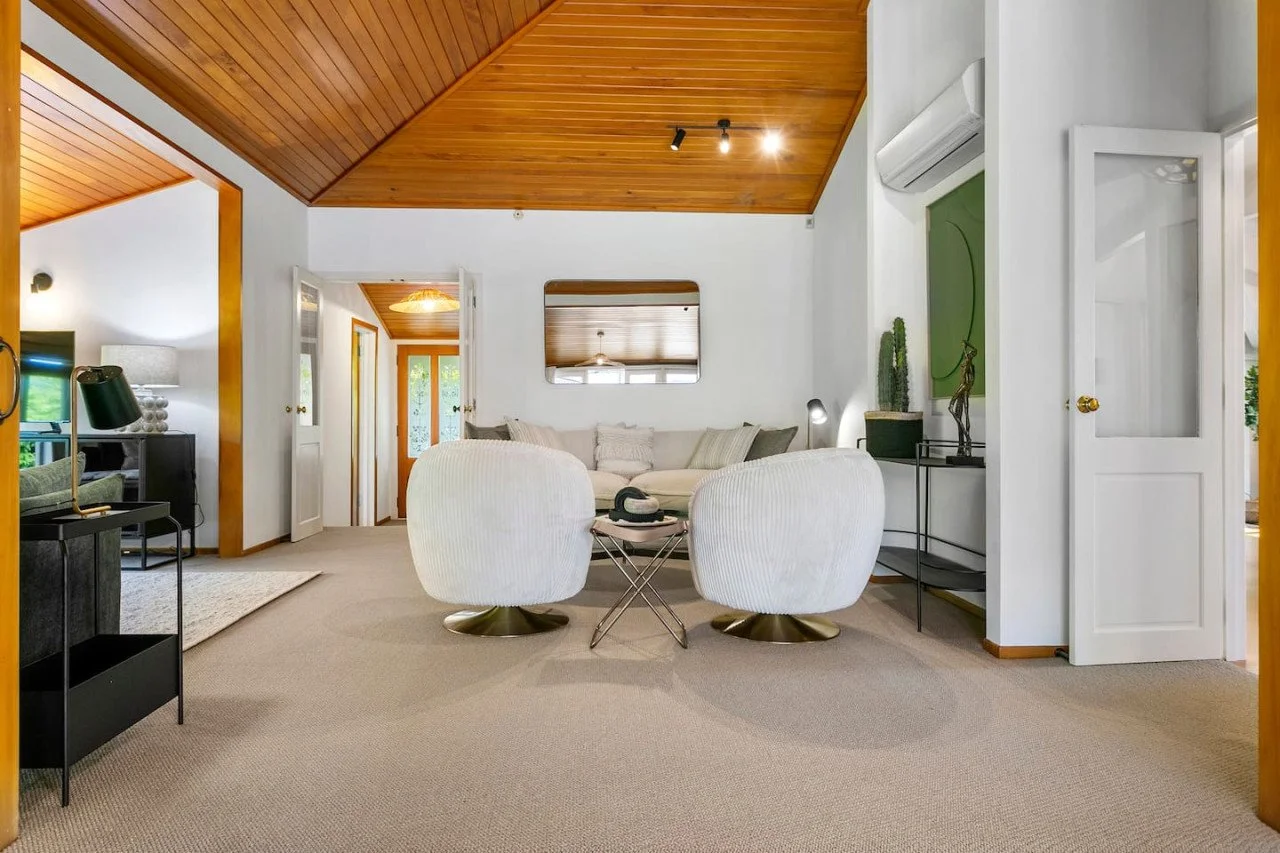 Living room with white couches, beige carpet, wood-paneled ceiling, green artwork, and various decorative items.