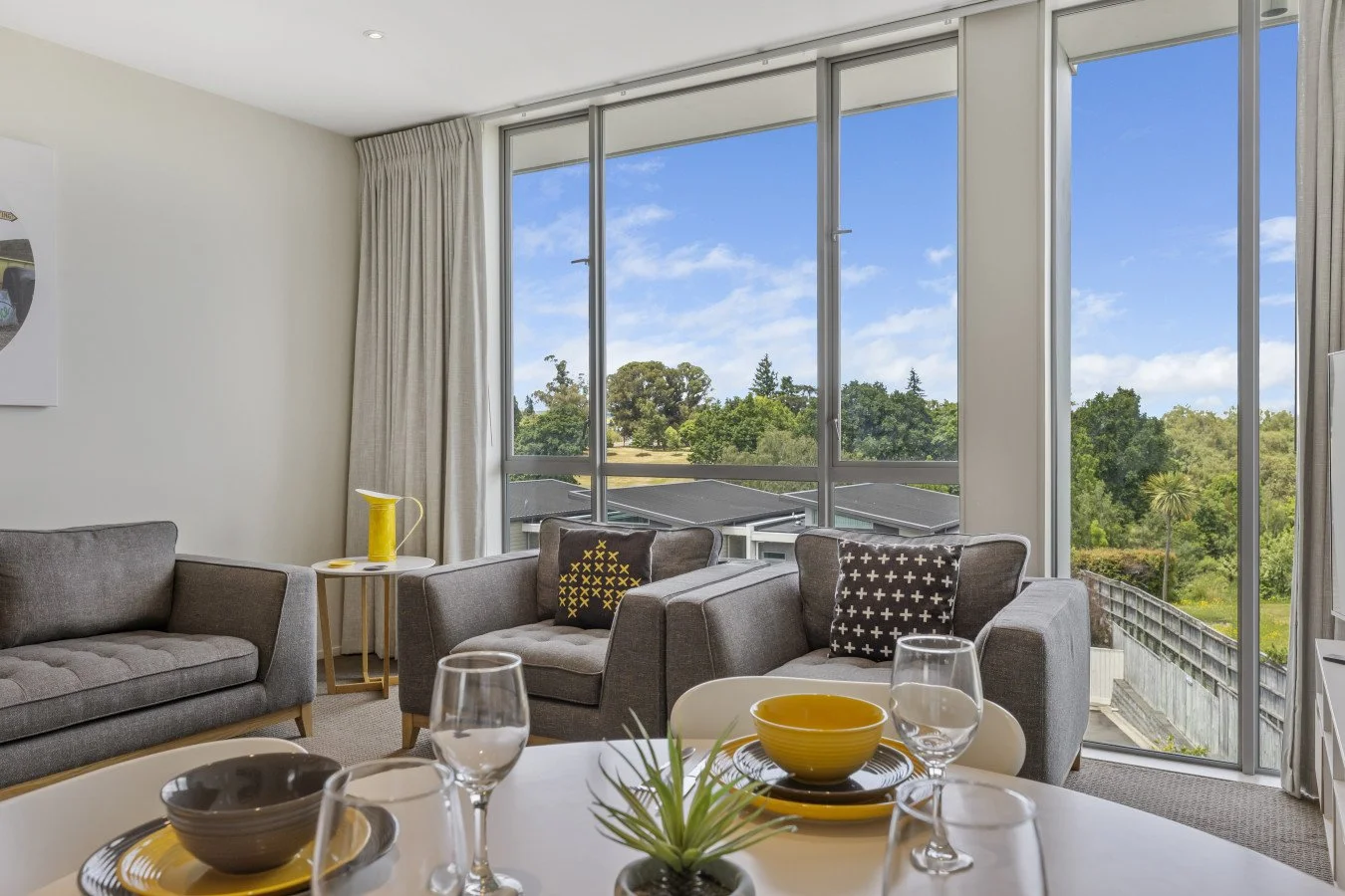 Living room with large windows showing trees outside, gray sofa, armchairs with black and yellow or black and white patterned pillows, a white dining table set with bowls, glasses, and a small plant, and a light-colored curtain.