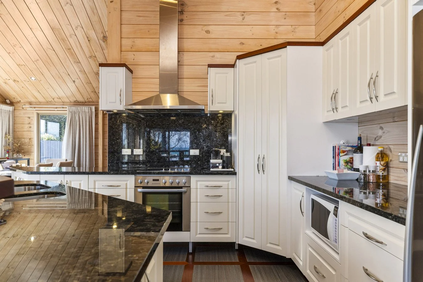 Modern kitchen with white cabinetry, black granite countertops, stainless steel oven and range hood, and wood-paneled walls and ceiling.