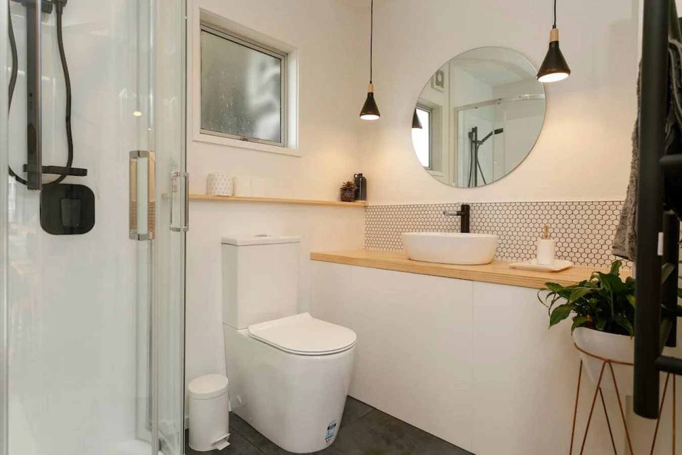 Modern bathroom with a toilet, a wooden vanity with a vessel sink, a round mirror, hanging pendant lights, a potted plant, and a shower enclosure with glass door.