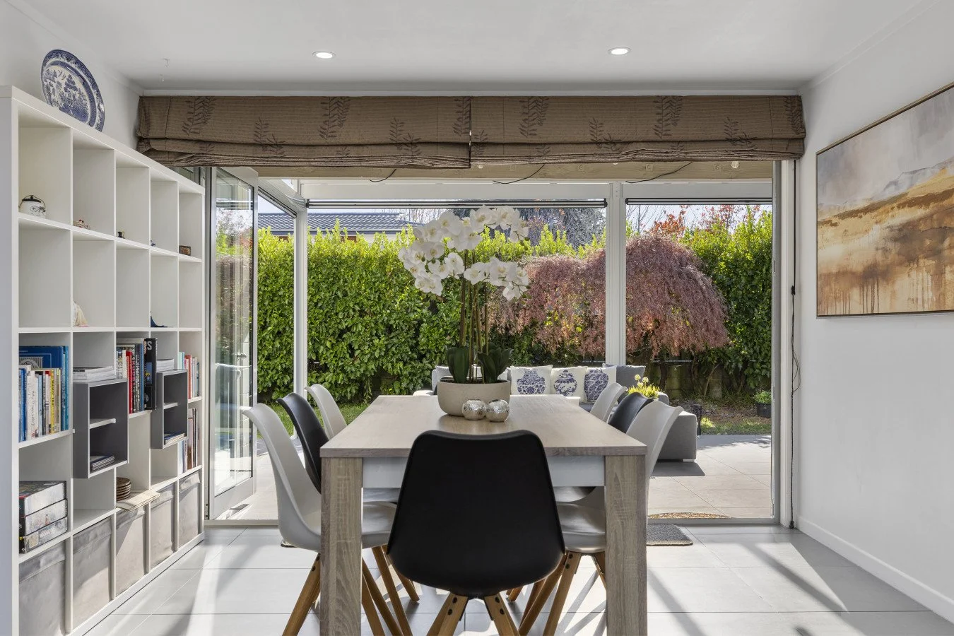 Dining room with a large wooden dining table, white and black chairs, a white orchid centerpiece, open sliding glass doors to a garden with green and purple foliage, white bookshelf on the left, abstract painting on the right wall, and natural light.