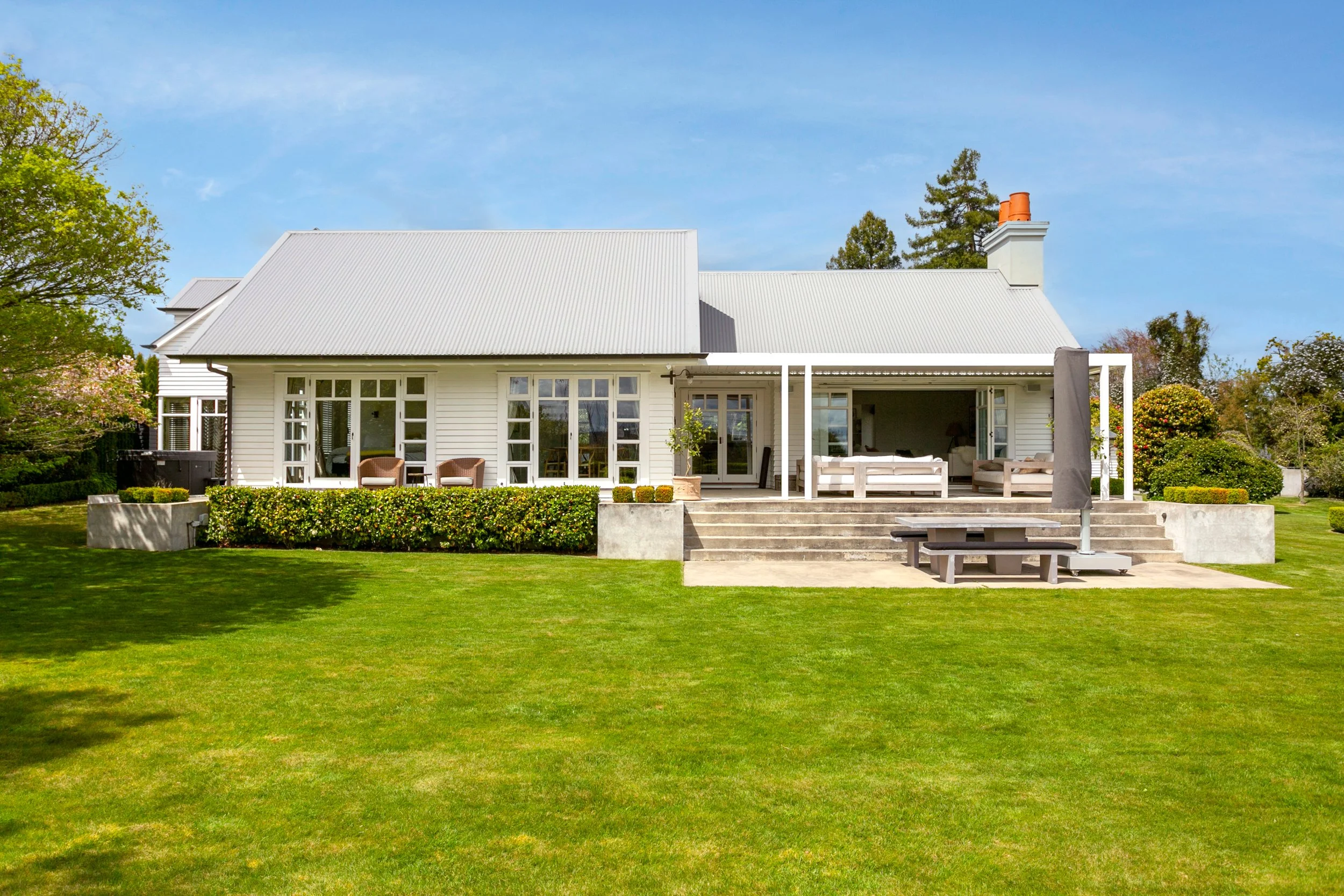 White house with a large front porch, outdoor seating, stairs, a picnic table with an umbrella, surrounded by green lawn and trees under a blue sky.