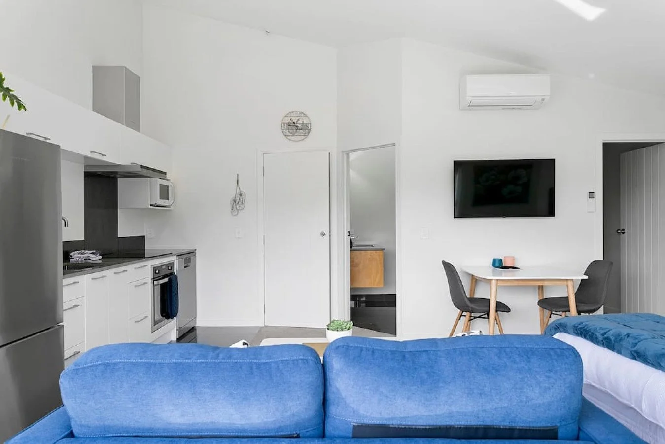 Modern living room and kitchen area with white walls, a blue sofa, a wall-mounted flat-screen TV, a small dining table with two chairs, and kitchen appliances including a refrigerator, microwave, and oven.