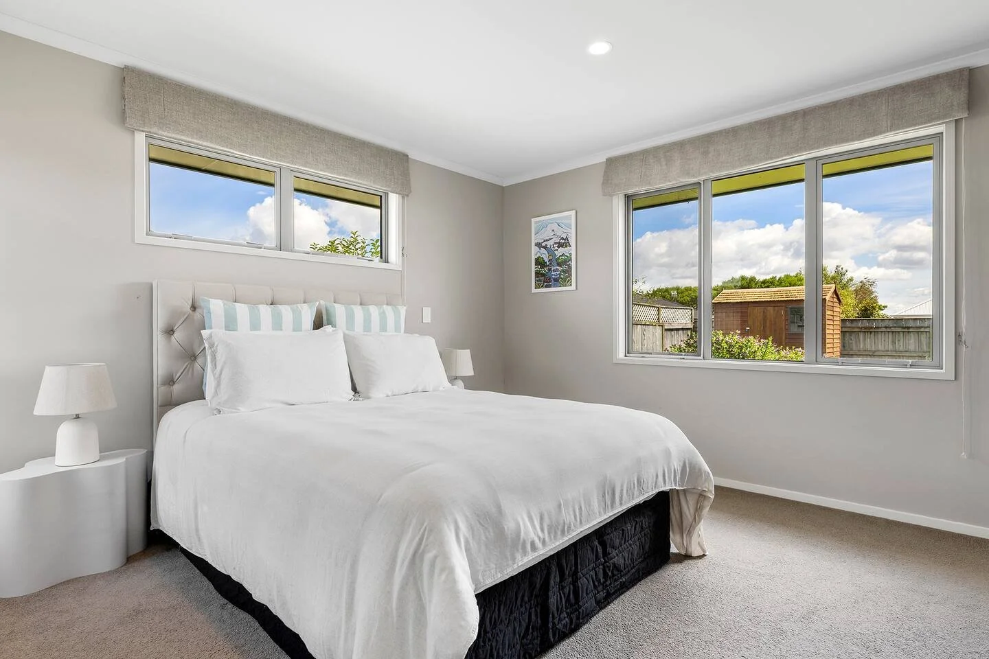 Bright bedroom with large windows, white bed with tufted headboard, white bedding, nightstand with white lamp, beige carpet, artwork on wall, view of a backyard with fence and shed outside.