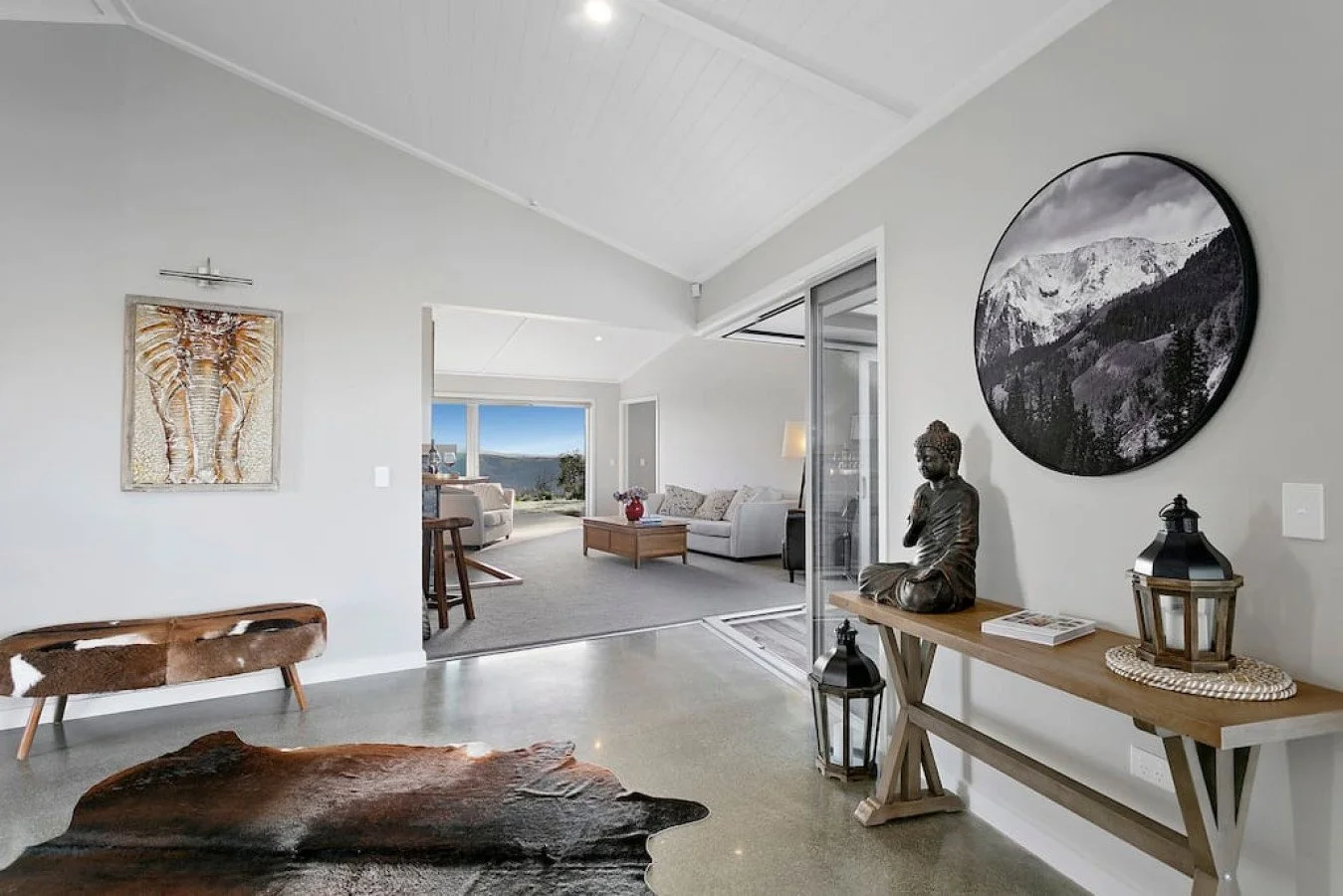 Living room with white walls, contemporary decor, a cowhide rug, artwork including an elephant painting and a mountain landscape photo, Buddha statue, lanterns, and a view of a porch with outdoor seating and distant mountains.