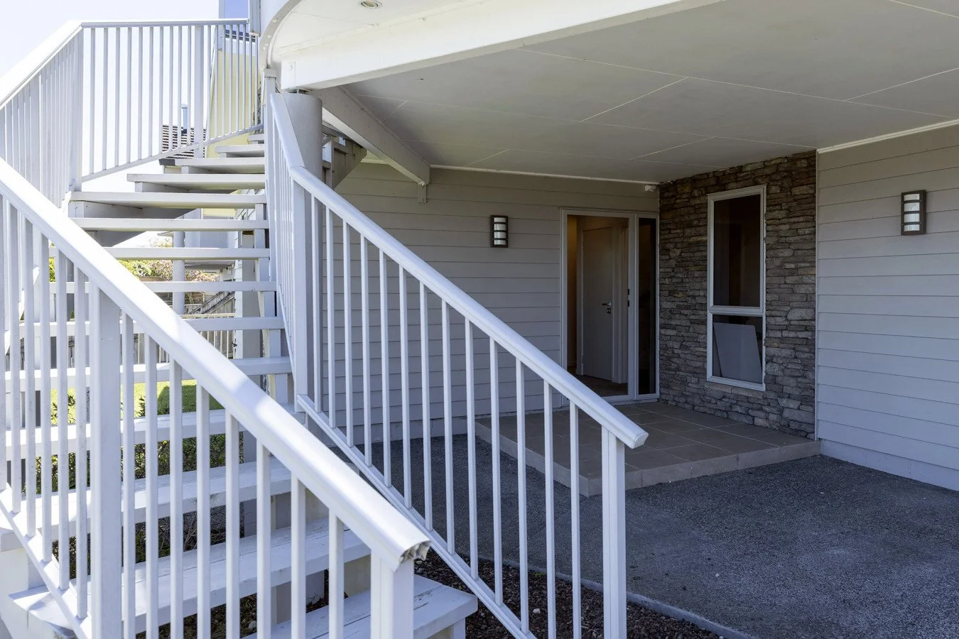 Exterior view of a modern house showing a white staircase with railing leading to an upper floor, a small porch with a concrete step, a sliding glass door, a window, and gray siding with a stone accent wall.