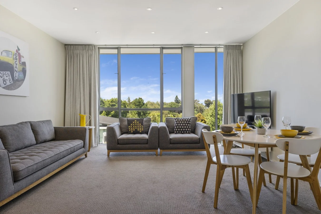 Living room with large window showing trees and blue sky, gray sofa, two gray armchairs, a round dining table set with bowls, plates, wine glasses, and a small plant, a flat-screen TV, and light-colored curtains.