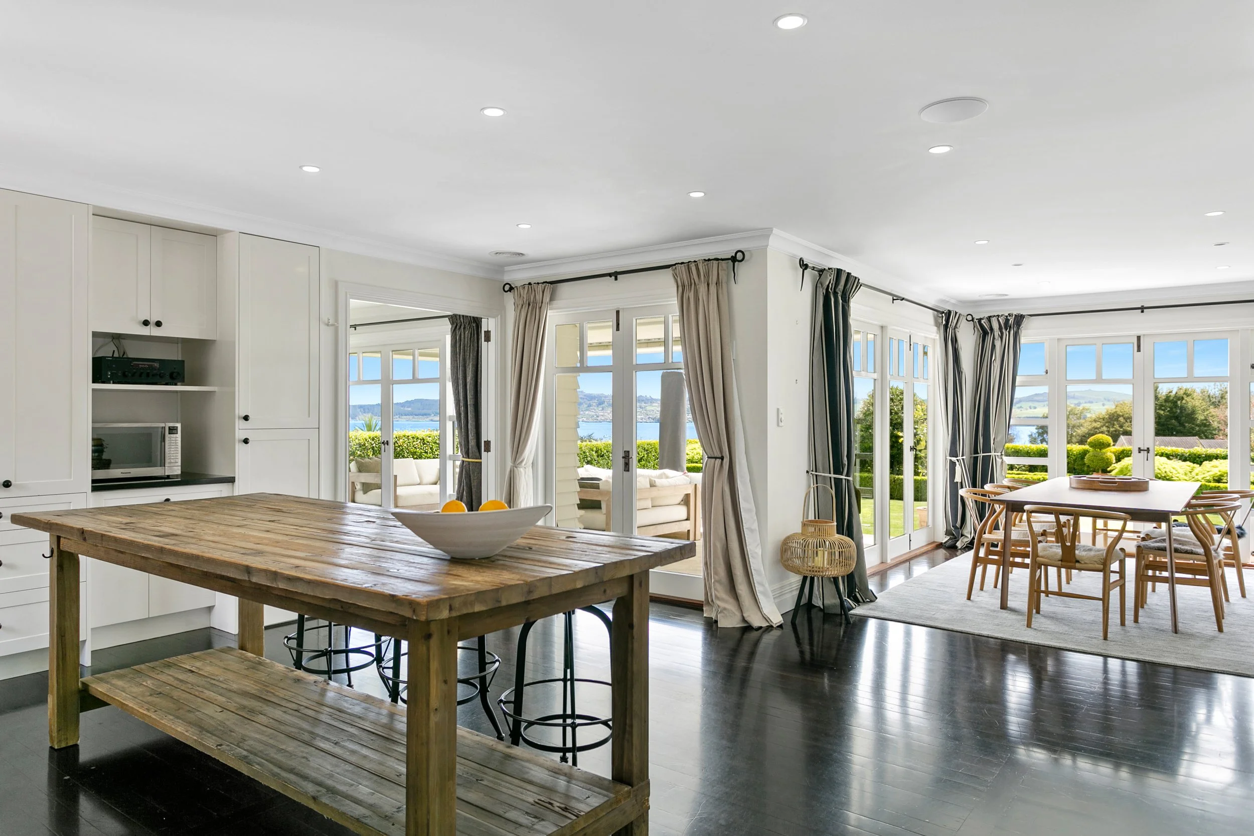 Bright, open kitchen and dining area with large windows and glass doors overlooking a garden and water view, furnished with a wooden island, chairs, and a dining table.