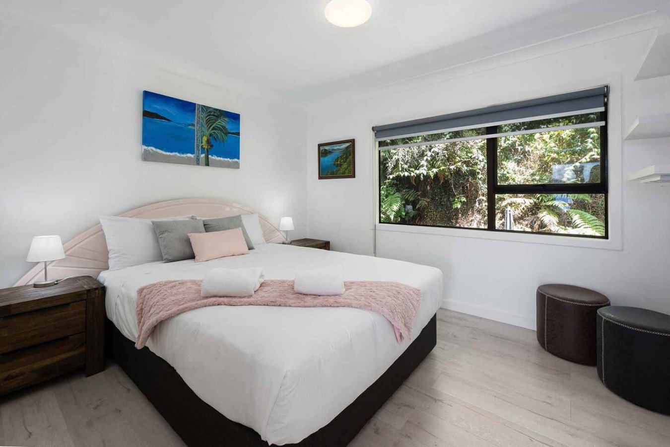 A bright bedroom with a large bed covered in white bedding, gray and pink pillows, and a pink throw blanket. There are wooden bedside tables with small white lamps on each side. A window with a view of green foliage and trees is on the right wall. Th