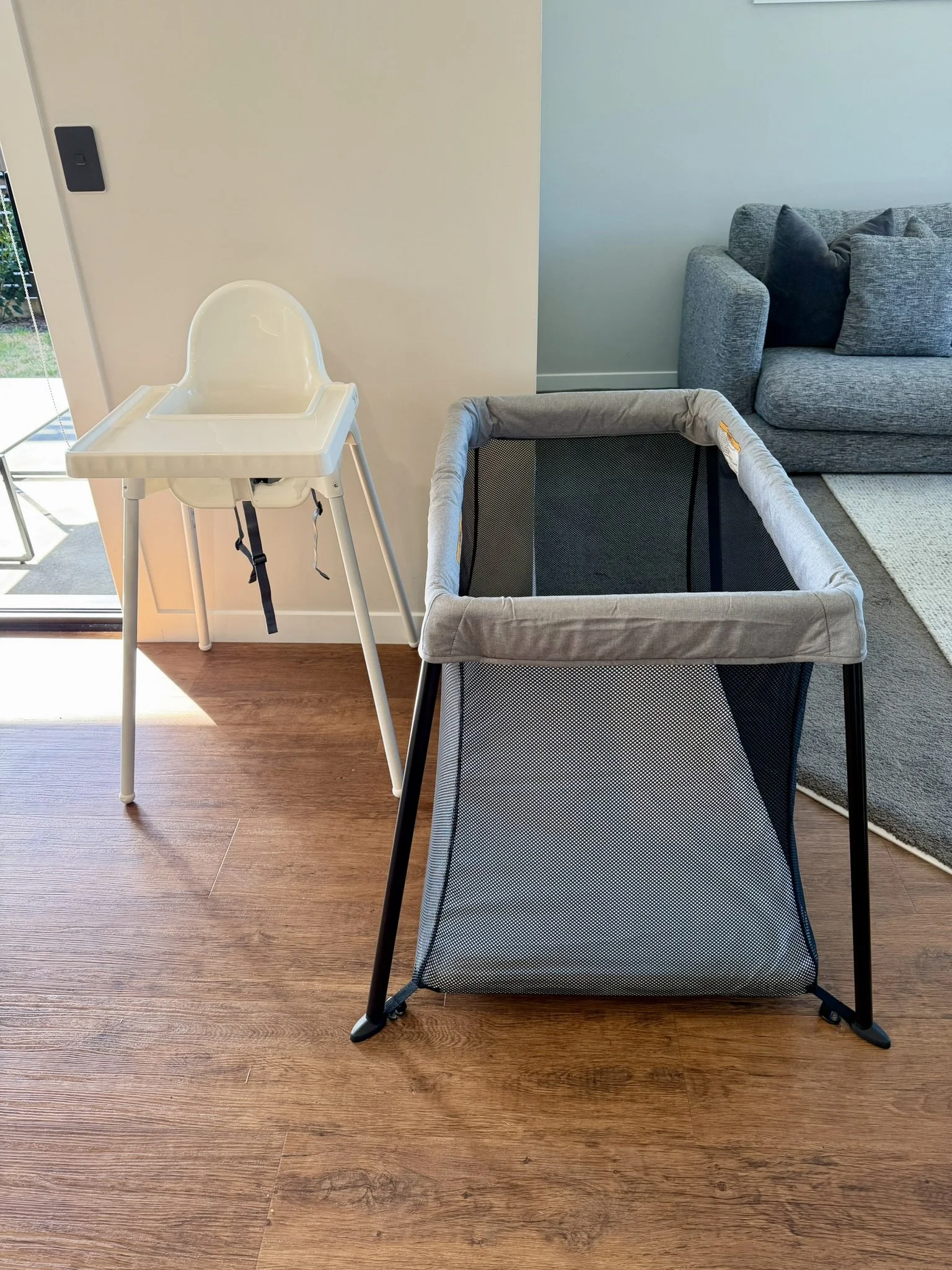 High chair and playpen in a living room with wooden flooring, gray sofa, and a green wall.