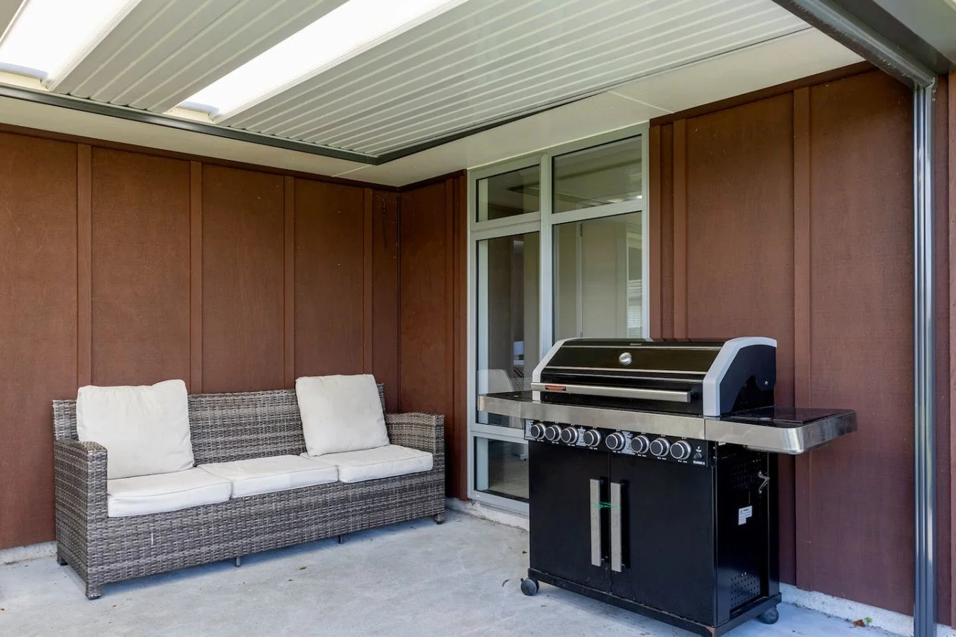 Covered outdoor patio with wicker sofa and white cushions, and a black gas grill, against brown wooden walls and glass door.