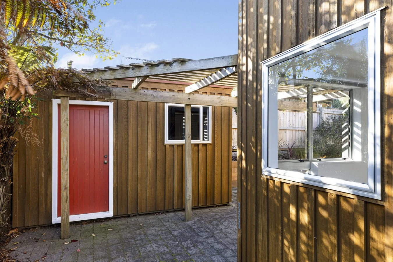 Exterior view of a small wooden shed with a red door, surrounded by a wooden fence and trees, on a paved patio.