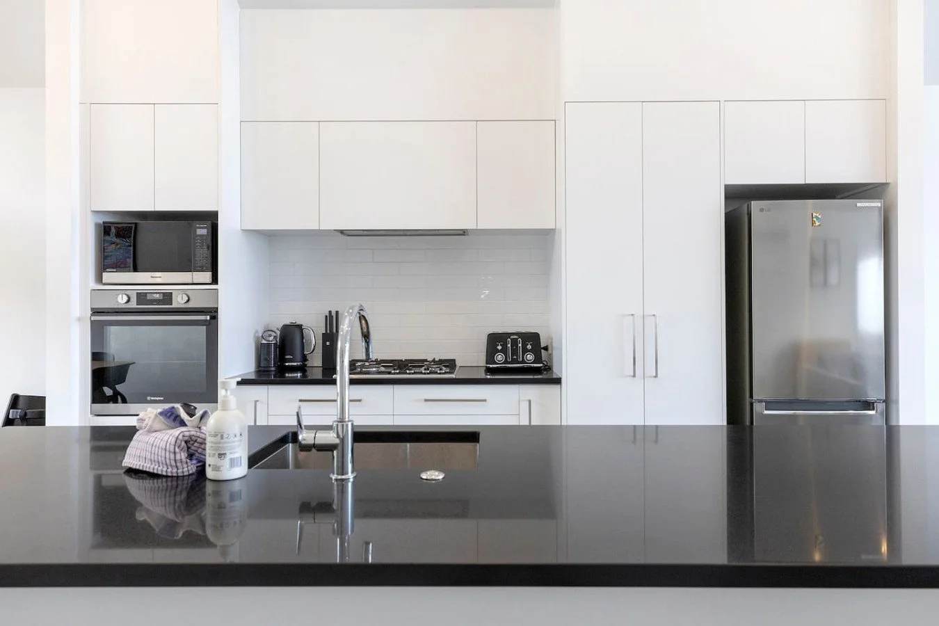 Modern kitchen with white cabinets, black countertop, and stainless steel appliances including a refrigerator, oven, microwave, and toaster, with a sink and soap dispenser on the island.