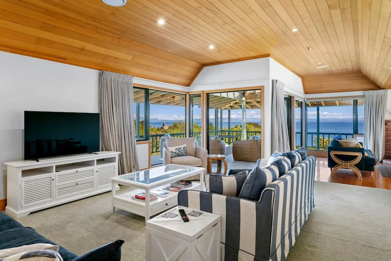 Living room with large windows showing ocean view, striped sofa, armchair, coffee table, TV, and balcony.
