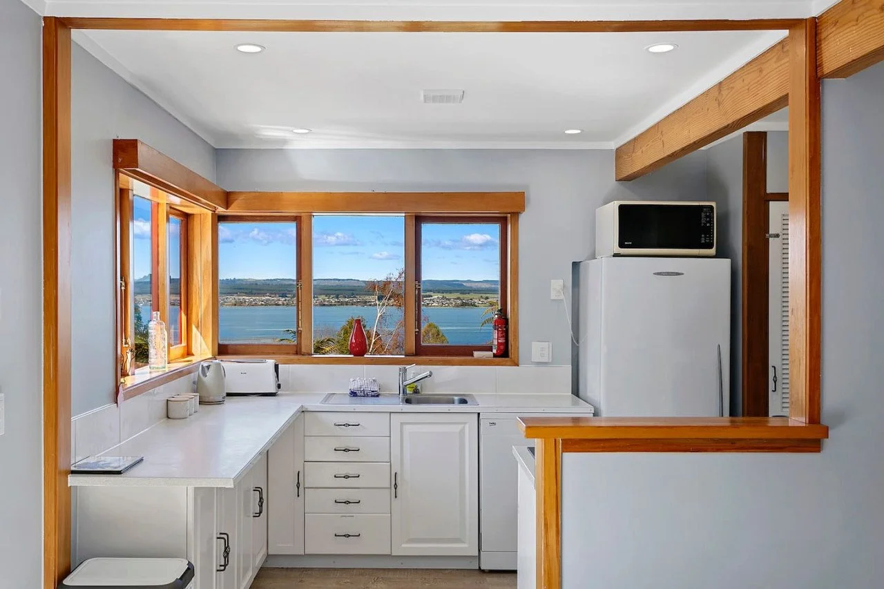 Kitchen with large window showing a scenic view of a lake and distant landscape, white cabinetry, microwave, refrigerator, countertops with small kitchen appliances, and wood trim accents.