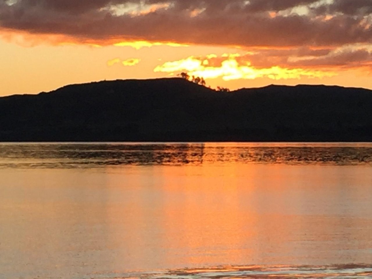 Sunset over a body of water with dark hills in the background, reflecting the orange and yellow colors of the sky.