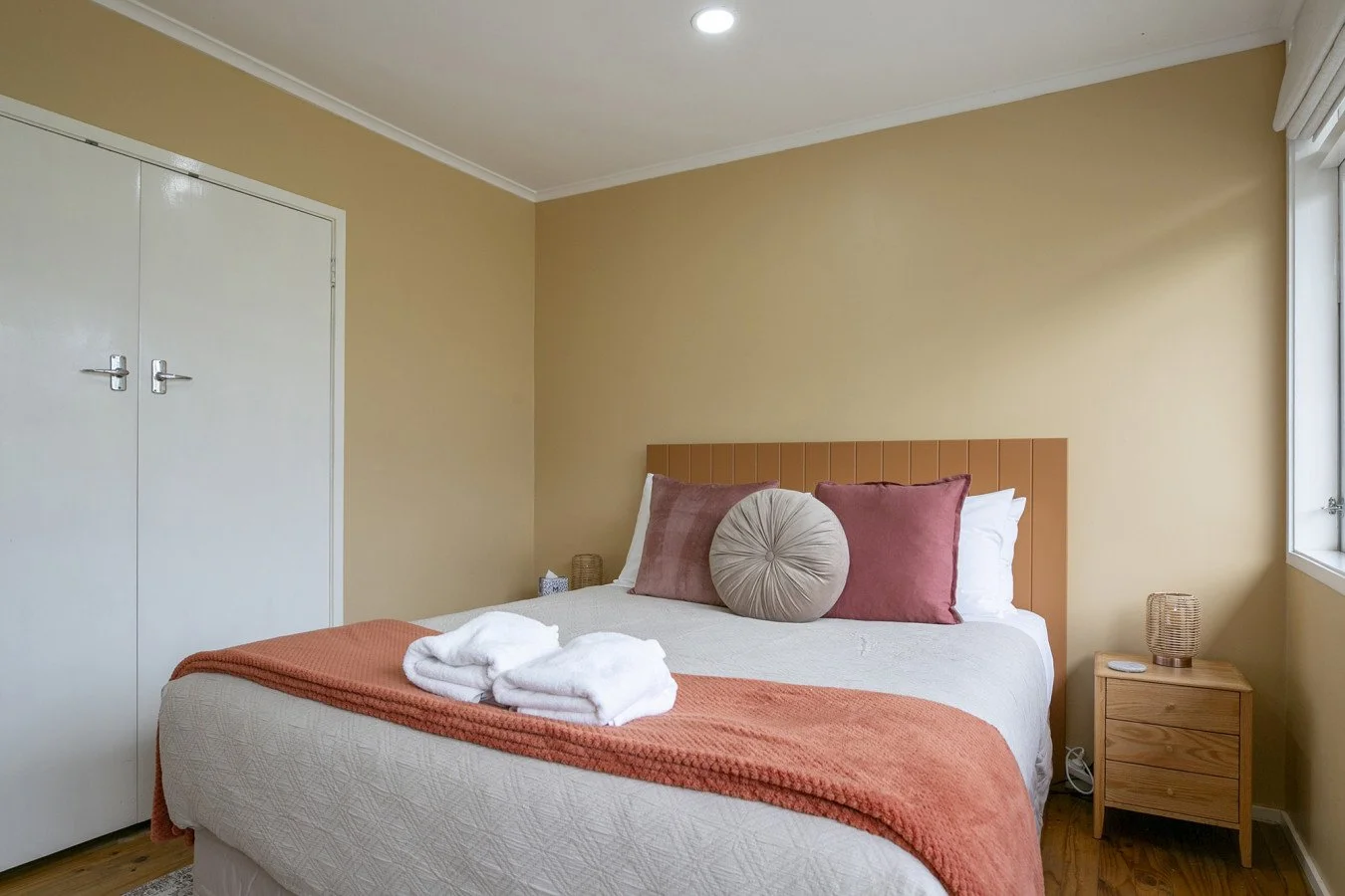 A bedroom with a bed, beige walls, a white closet door, a window, and a nightstand with a lamp.