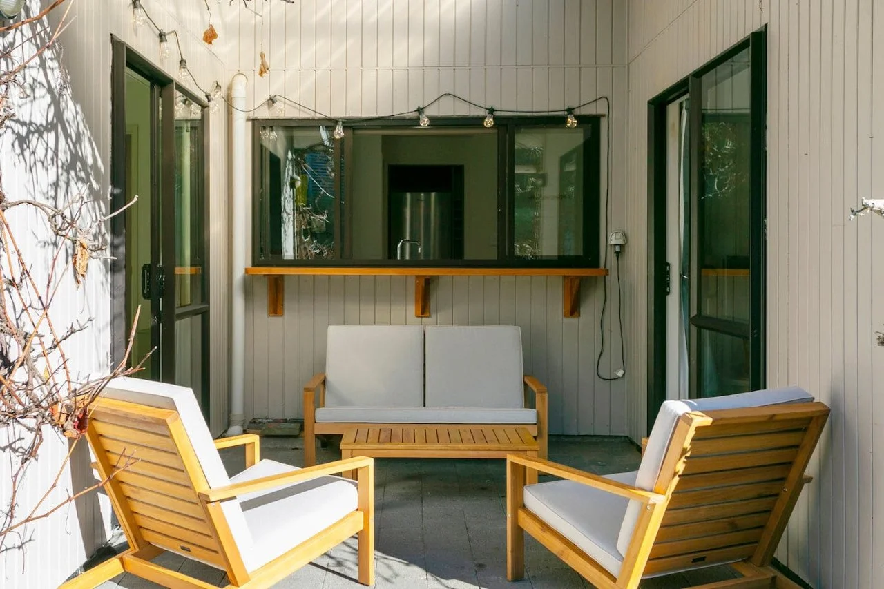 Outdoor patio with wooden furniture including a loveseat and two armchairs with white cushions, a wall with large windows, string lights, and a small bar-height table attached to the wall.