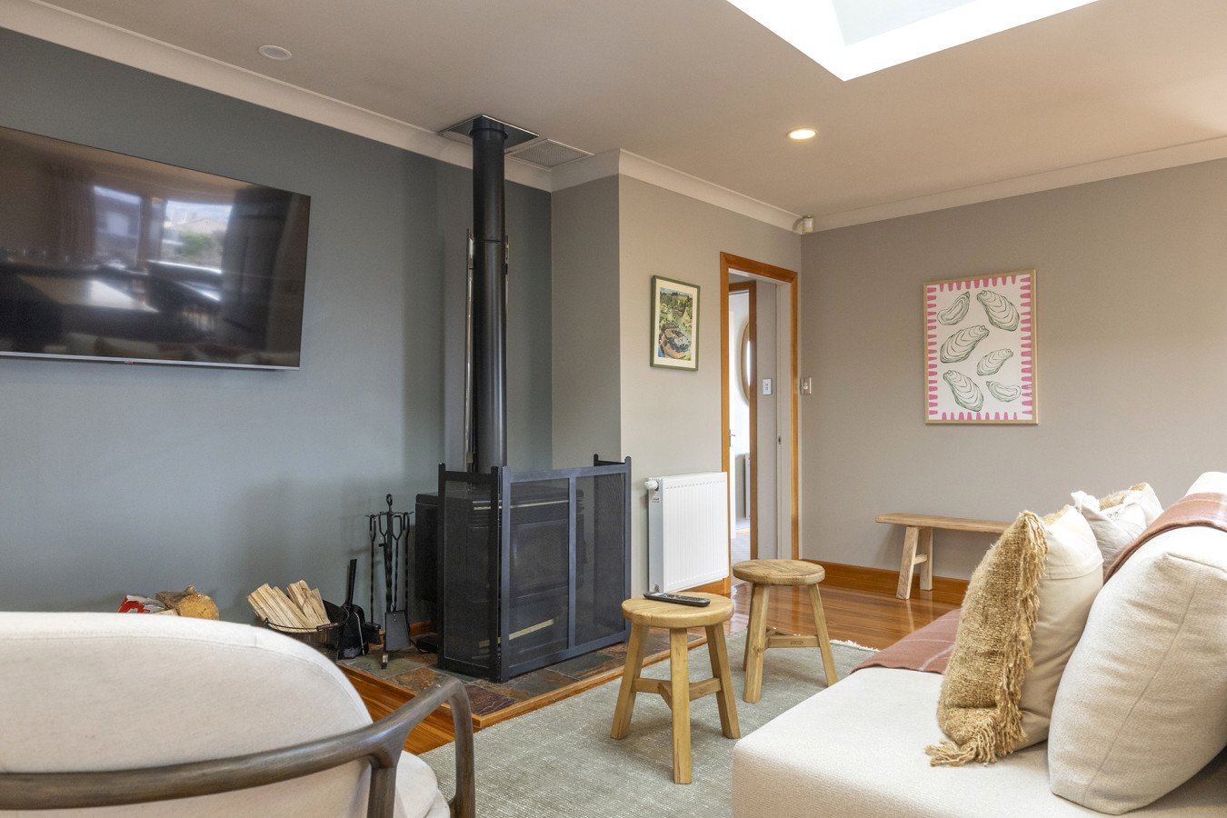Living room with gray walls, a mounted flat-screen TV, a black wood stove with fireplace tools, two small wooden stools, a beige sofa with decorative pillows, framed artwork on the walls, a bench, and hardwood flooring.