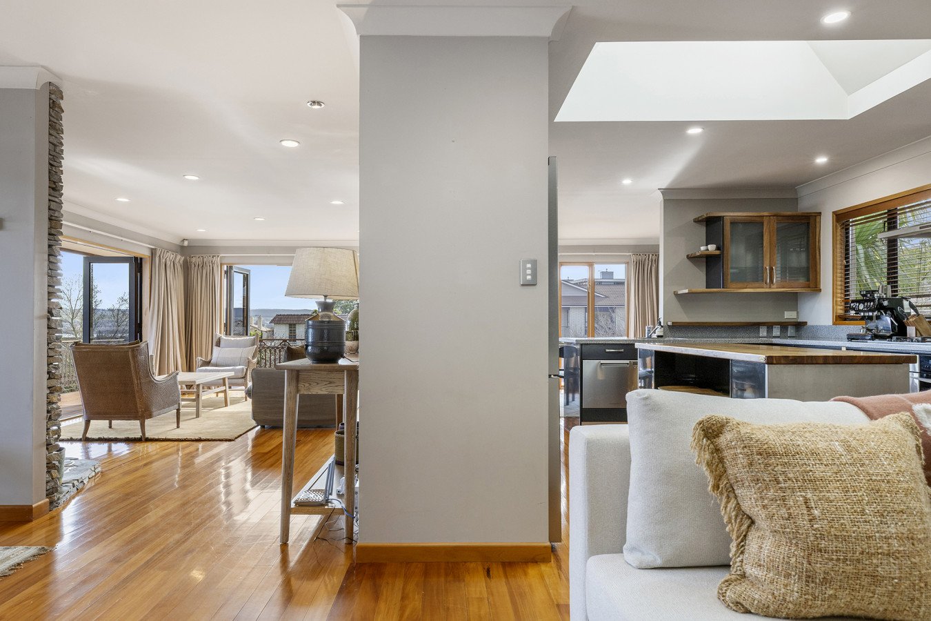Open-concept living room and kitchen with hardwood floors, large windows, neutral-colored furniture, and a kitchen with wooden accents and black appliances.