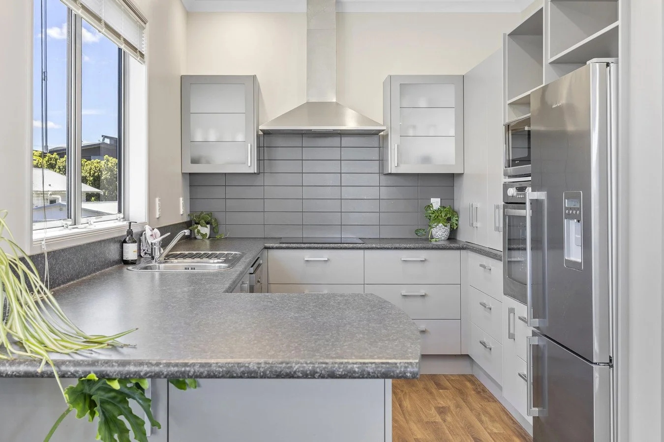 Modern kitchen with gray cabinets, stainless steel appliances, gray backsplash tiles, and wooden flooring, with a large window above the sink.