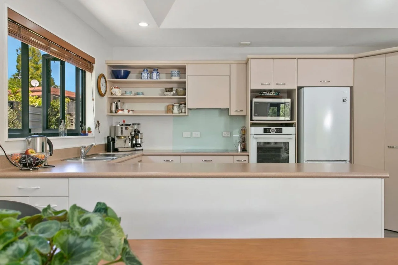 Modern kitchen with beige cabinets, a window with bamboo blinds, and a countertop with a fruit basket and coffee machine.