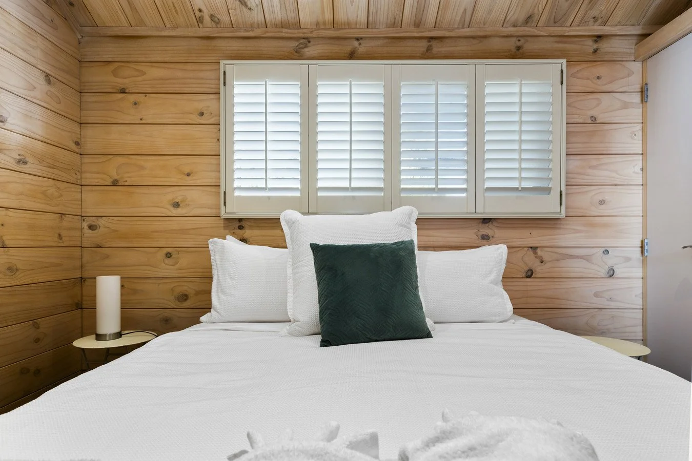 A cozy bedroom with wood-paneled walls, a white bed with pillows, and a green accent pillow. A window with white shutters is above the bed, and small bedside tables with lamps are on either side.