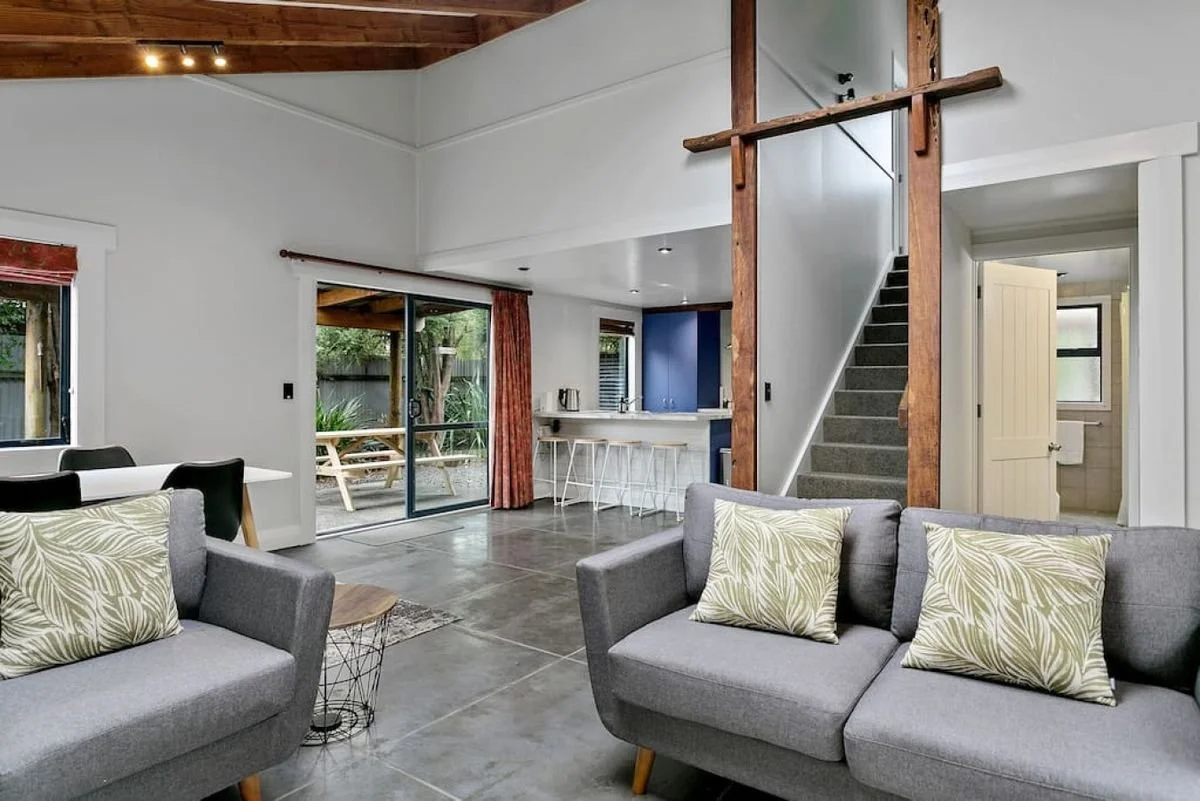 Living room with two gray sofas with green and white patterned pillows, a glass sliding door leading outside, a small round side table, a dining area with a white table and black chairs, a kitchen with a blue accent wall, barstools, and a staircase w