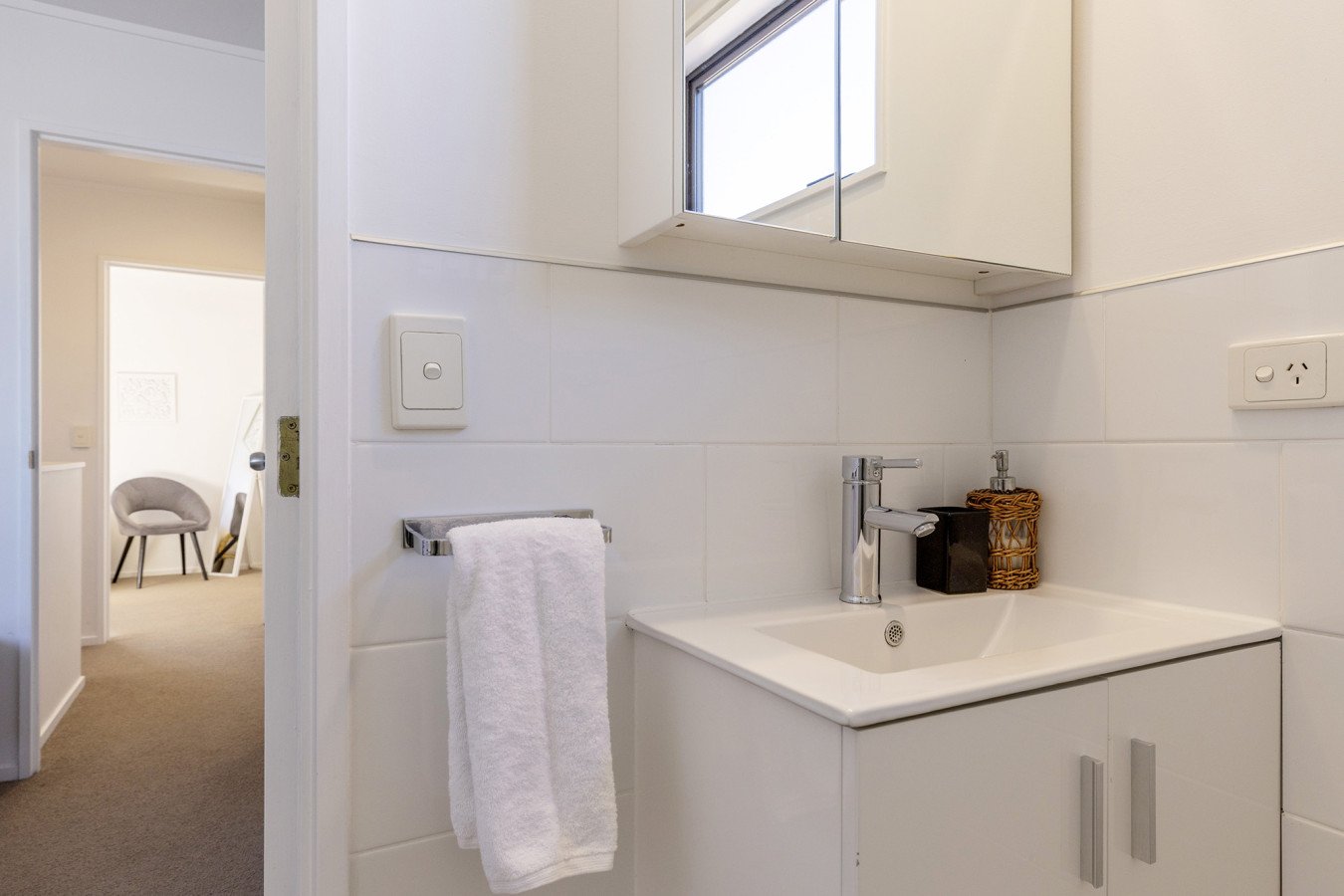 A small bathroom sink with a chrome faucet, a mirror cabinet above, and a window. There is a white towel hanging on a towel rack next to the sink, and a soap dispenser and a container are on the sink. The walls are tiled white.