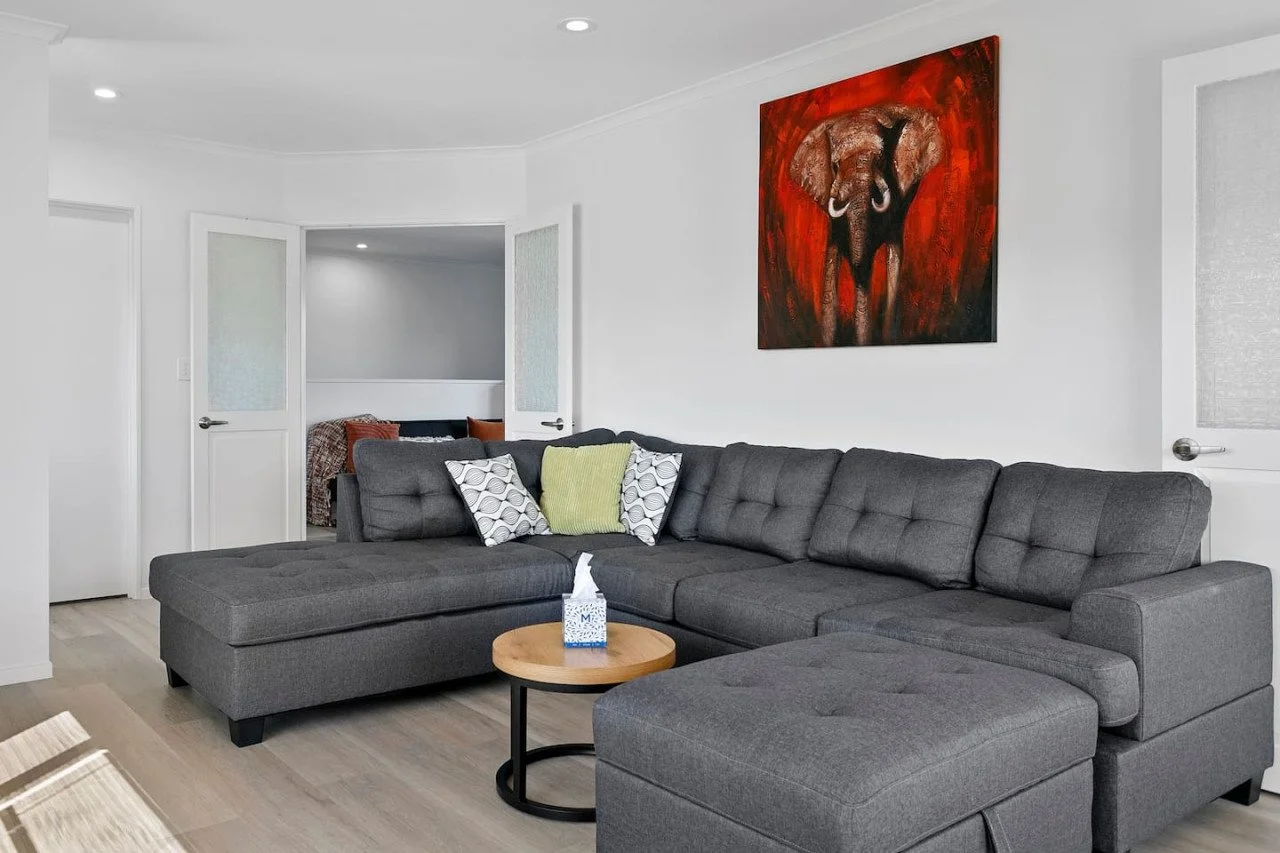 Modern living room with white walls and gray sectional sofa decorated with patterned and solid-colored throw pillows, a small round wooden coffee table with tissues, and a red-toned elephant painting on the wall.