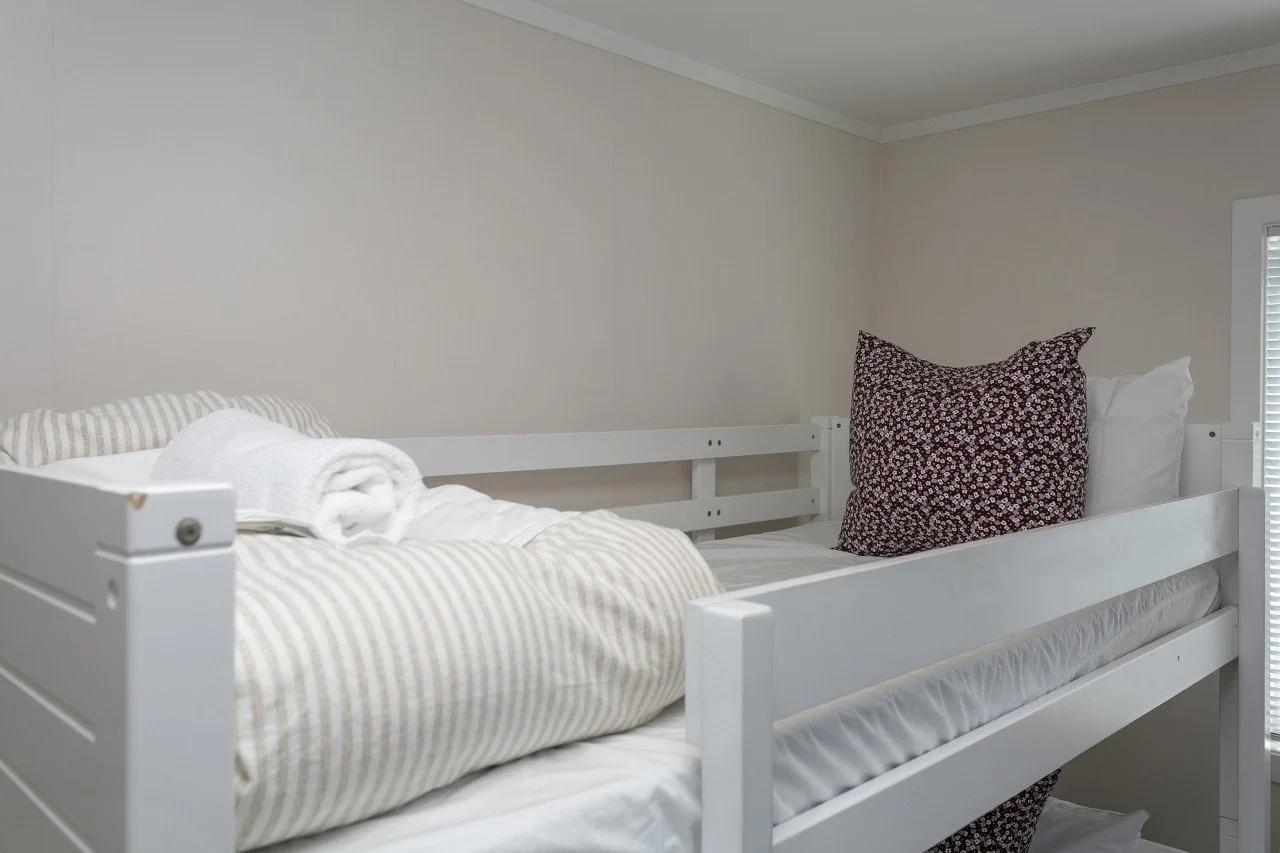 A neatly made bunk bed with white frames, striped beige and white bedding, a rolled white towel, and patterned pillows against a light-colored wall.