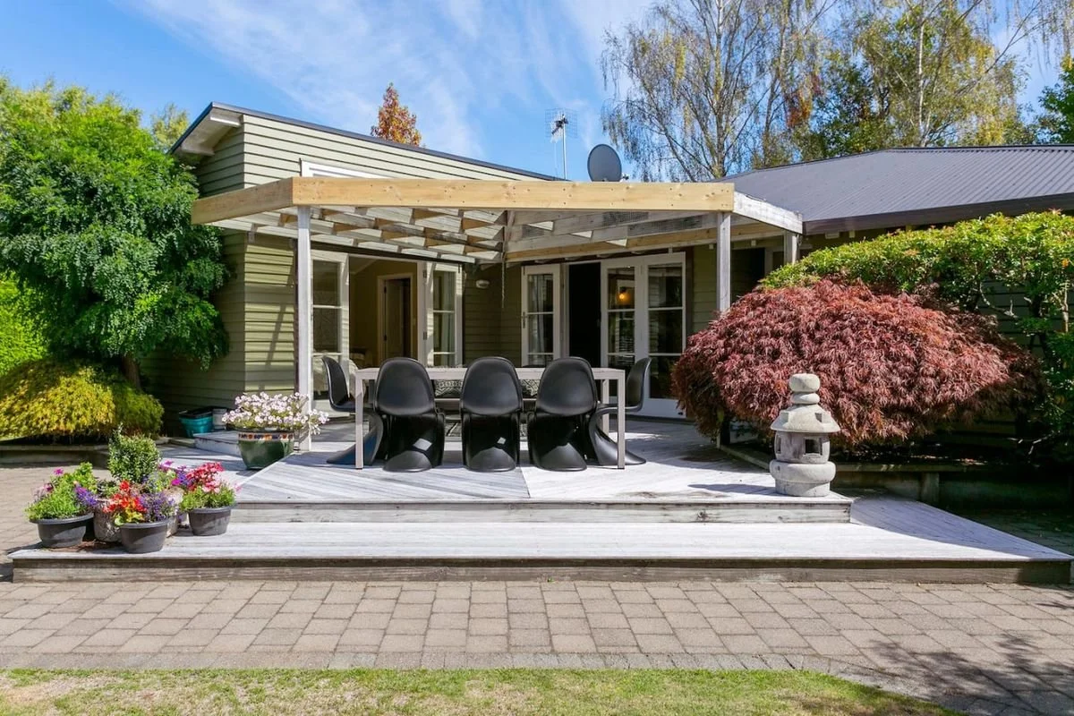 A house with a wooden deck, black chairs around a dining table, and potted plants and flowers on the deck. There is a Japanese stone lantern and a Japanese maple tree in front.