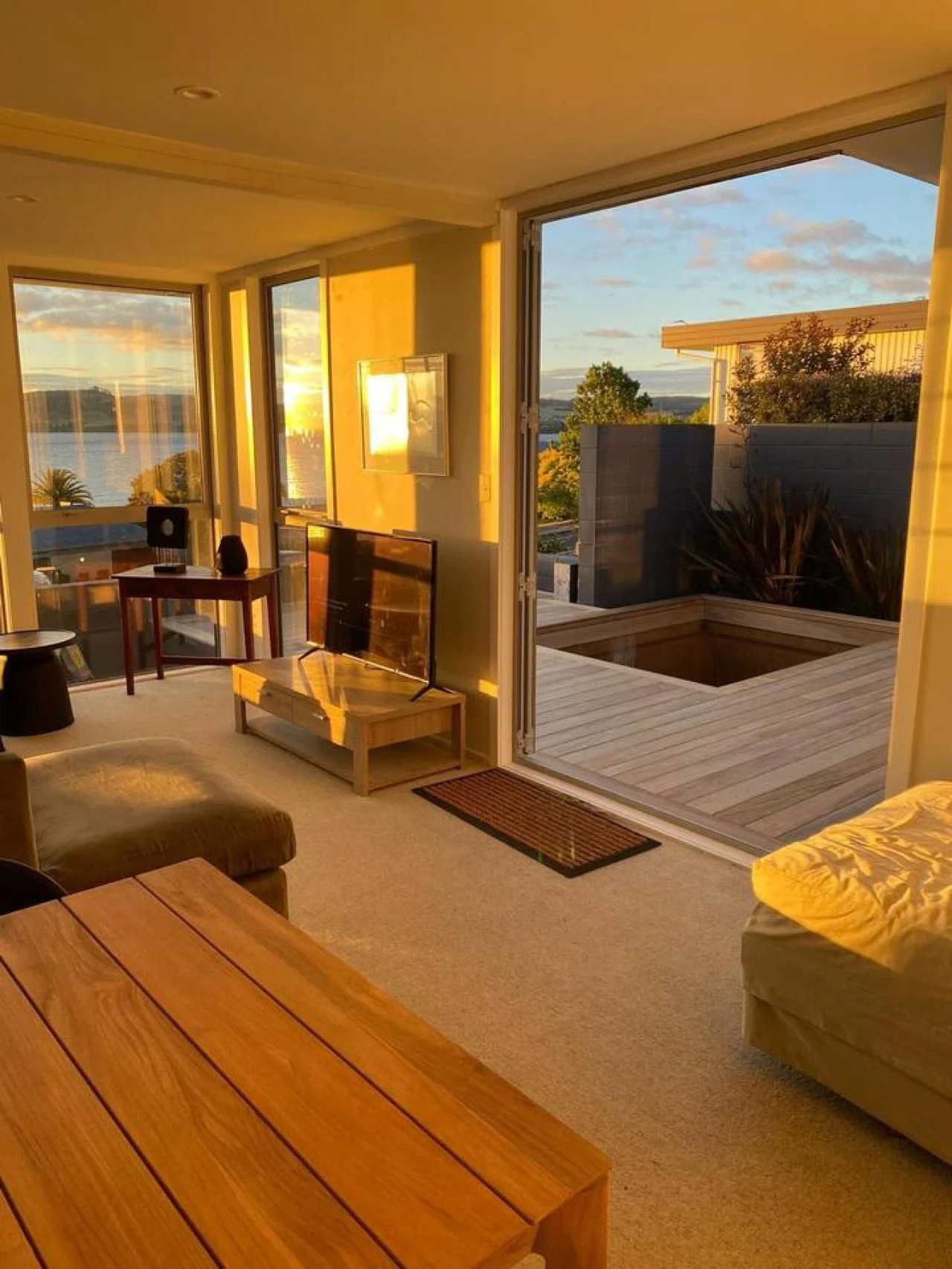 Living room at sunset with large windows, a television, and outdoor deck with hot tub, overlooking a body of water and trees.
