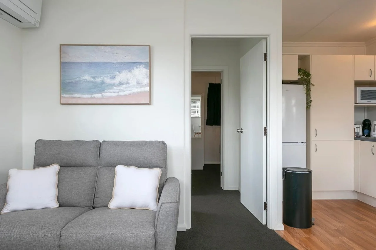 Living room with a gray sofa and white pillows, a wall art of a beach with waves, and a view into a kitchen and a small room with a window and dark curtains.
