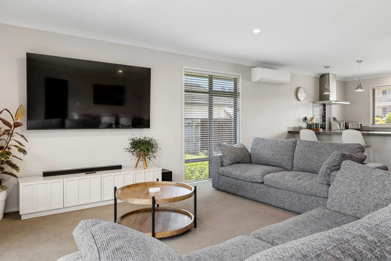 Living room with a large wall-mounted TV, a gray sofa, a round wooden coffee table, a white entertainment unit, and a window with blinds, with visible kitchen area in the background.