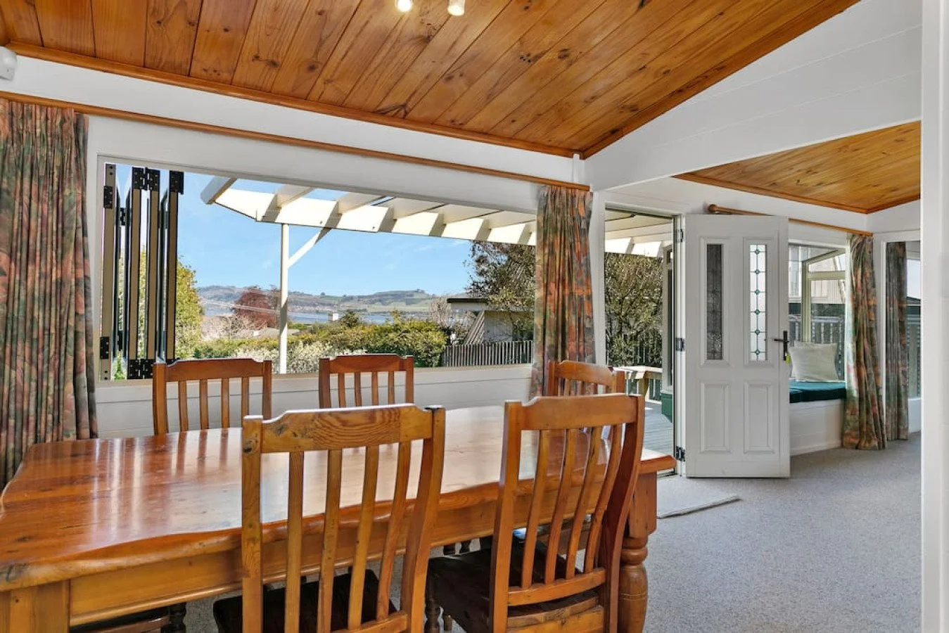 A dining room with a wooden table and six matching chairs, large windows with floral curtains, and a door leading to a porch with a view of a landscape with trees and hills.