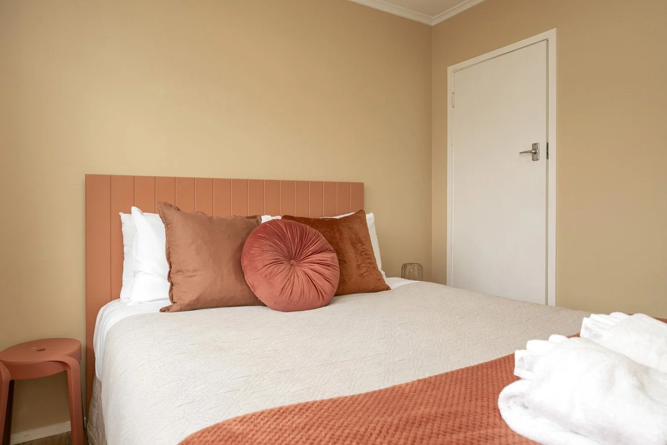 A neatly made bed with white bedding, multiple brown and rust-colored pillows, and a rust-colored blanket at the foot of the bed. A peach-colored headboard is on the bed, and a small peach-colored side table sits beside it. A white door is on the bei