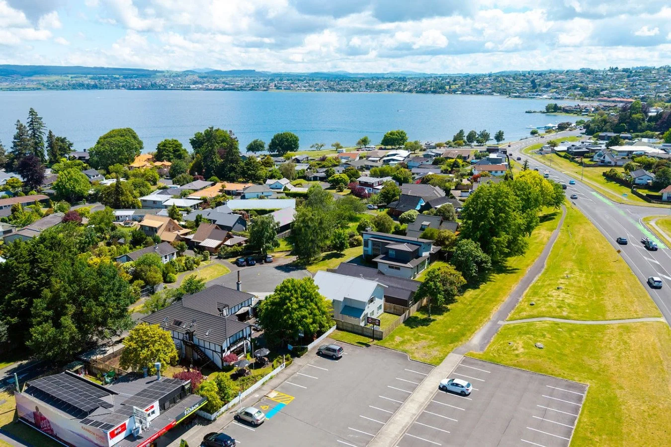 Aerial view of a lakeside residential area with numerous houses, green trees, a parking lot, a road, and a large body of water in the background, under partly cloudy skies.