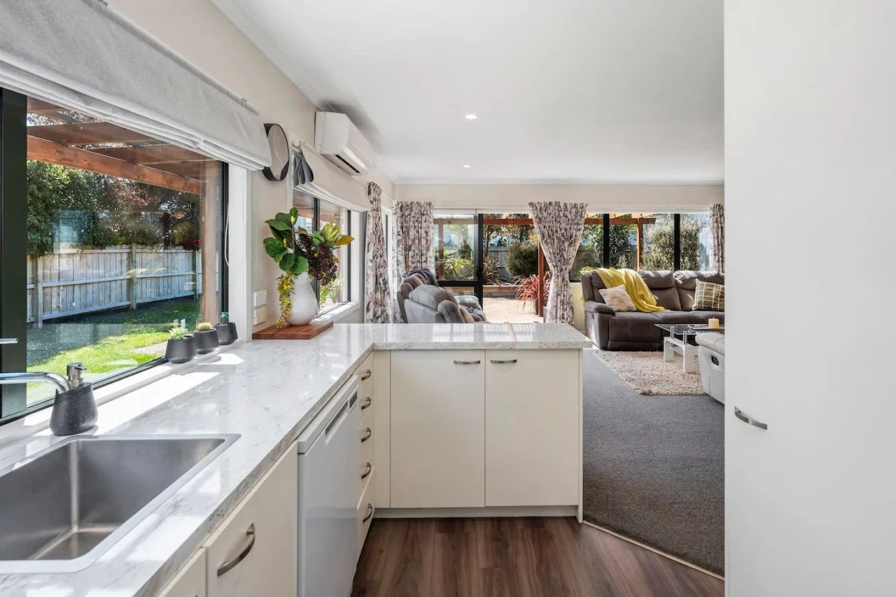 A view from the kitchen looking into a bright living room with large windows, curtains, sofas, and patio doors, with outdoor greenery visible.