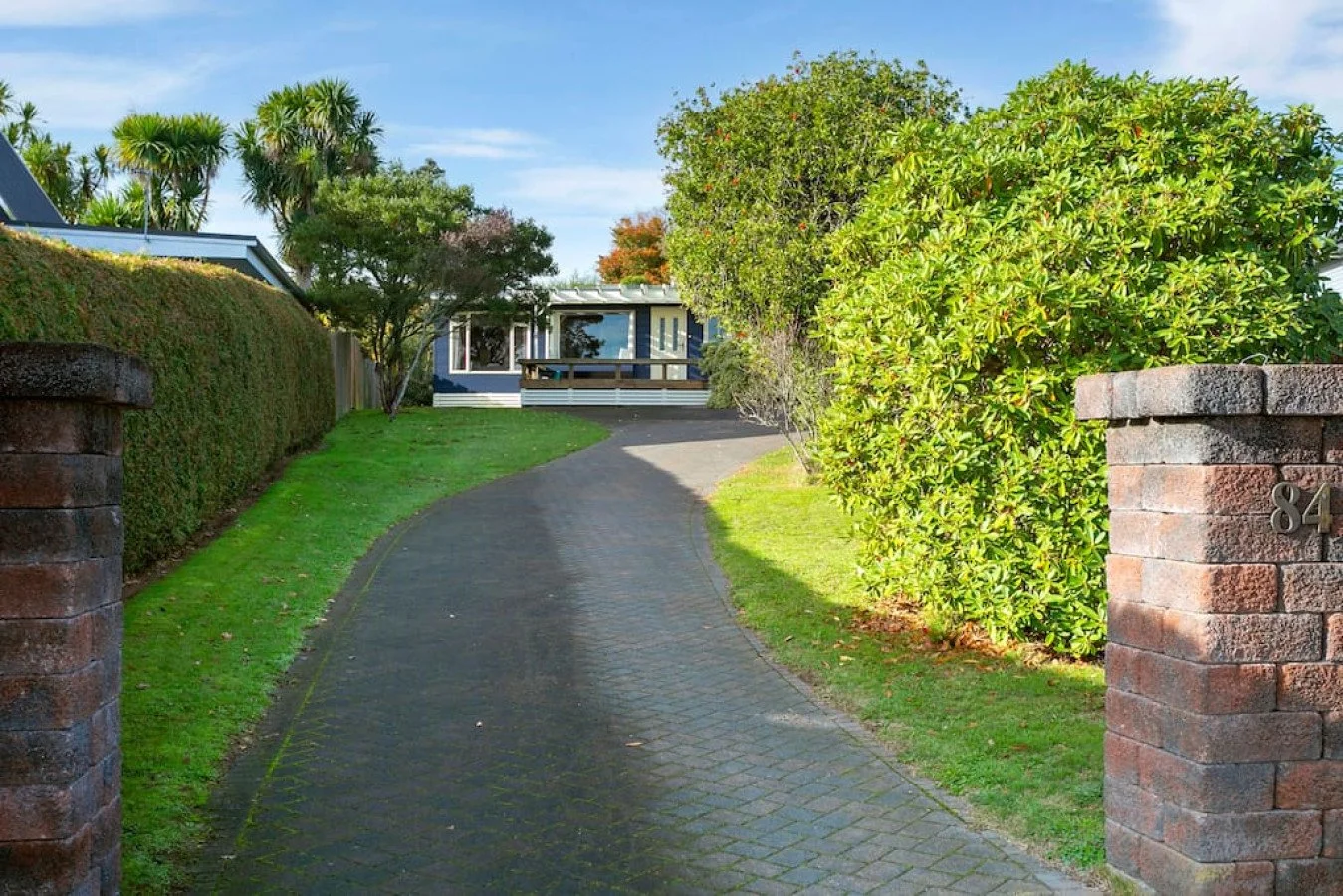 Paved driveway leading to a house with a blue exterior, surrounded by greenery, with brick pillars at the entrance marked with the number 84.