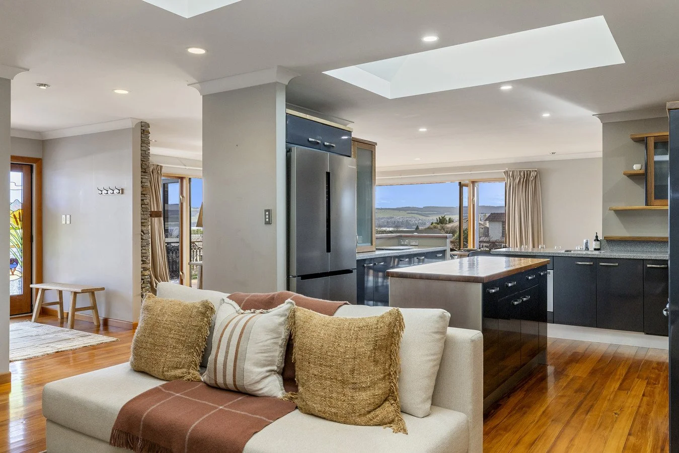 Open-concept living room and kitchen area with wooden floors, a white sofa with pillows, large windows, and a stainless steel refrigerator.
