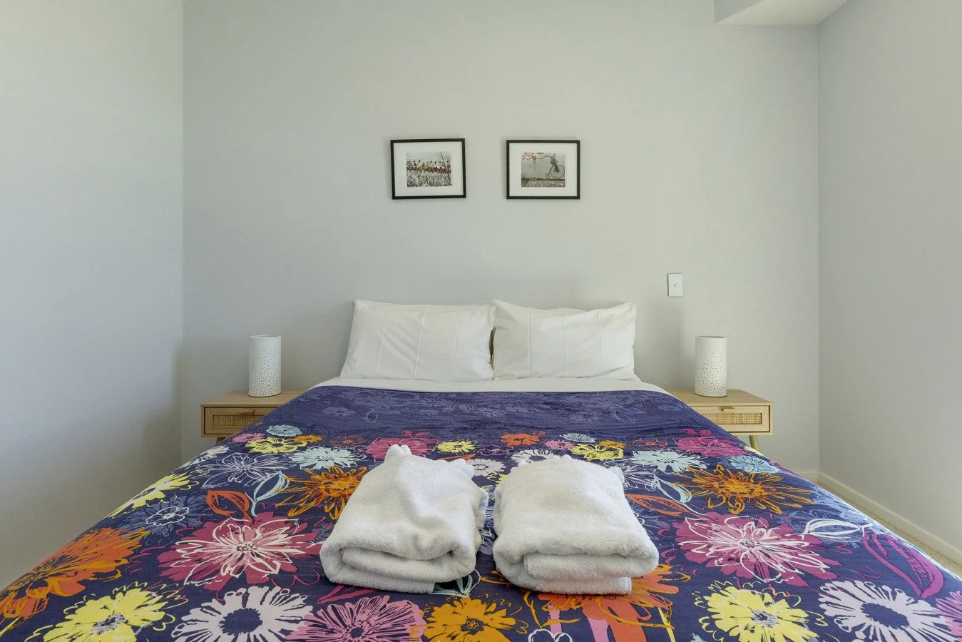 A bedroom with a bed featuring a colorful floral bedspread, two folded white towels on the bed, white pillows, two nightstands each with a white lamp, and two framed pictures on the wall above the bed.