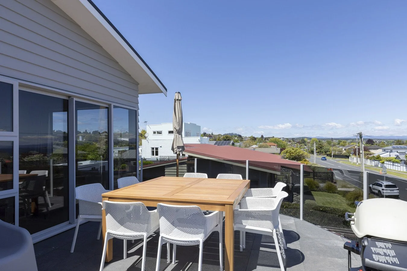 Outdoor patio with a wooden dining table, white chairs, and a closed patio umbrella, overlooking a neighborhood with houses, trees, and a street under a clear blue sky.