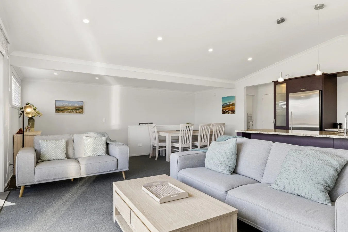 Living room with light-colored sofas, a wooden coffee table, and artwork on the walls; open kitchen with dark cabinetry and pendant lighting.