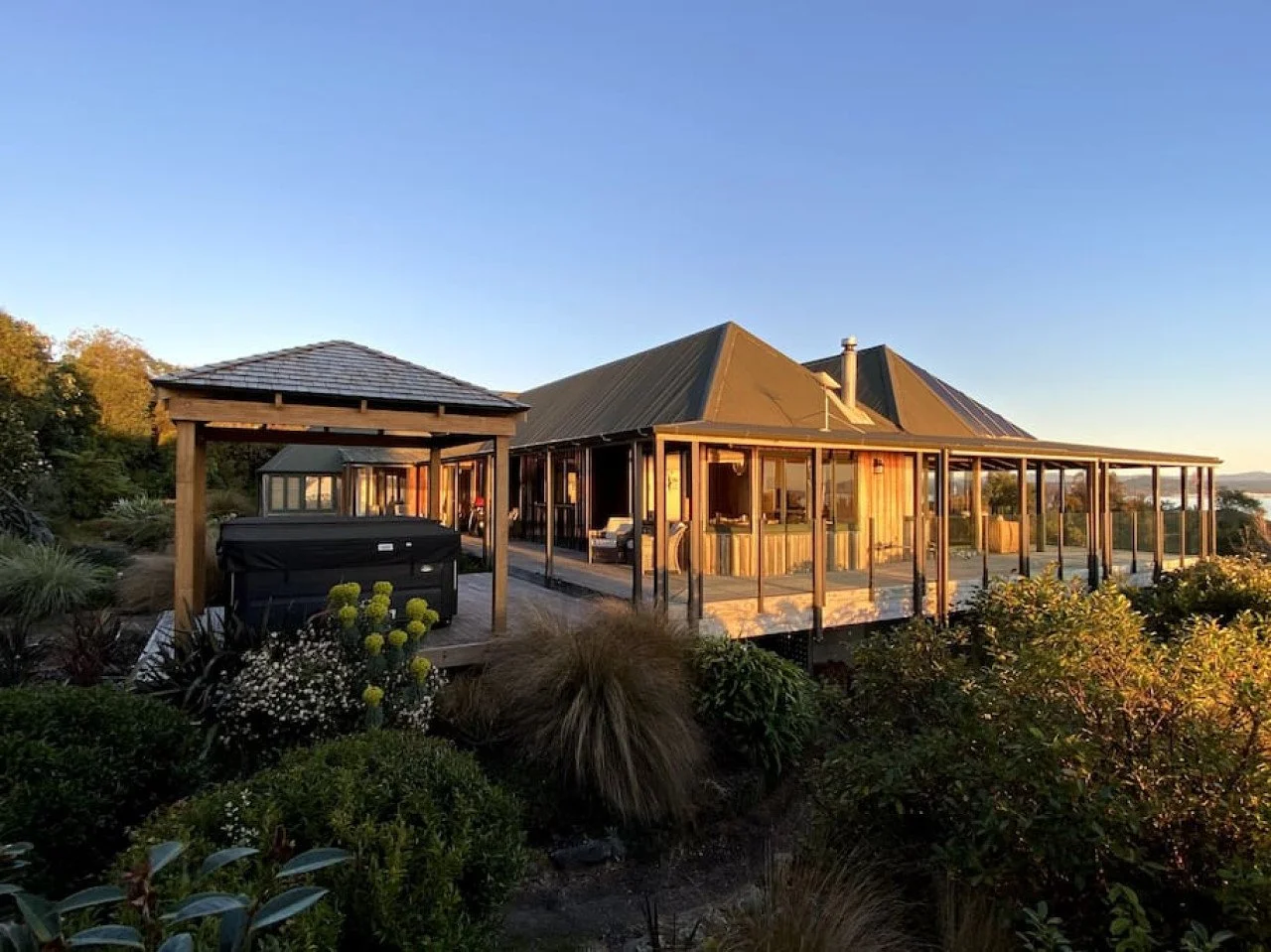 A wooden house with a metal roof and a wraparound porch, surrounded by greenery and plants, during sunset.