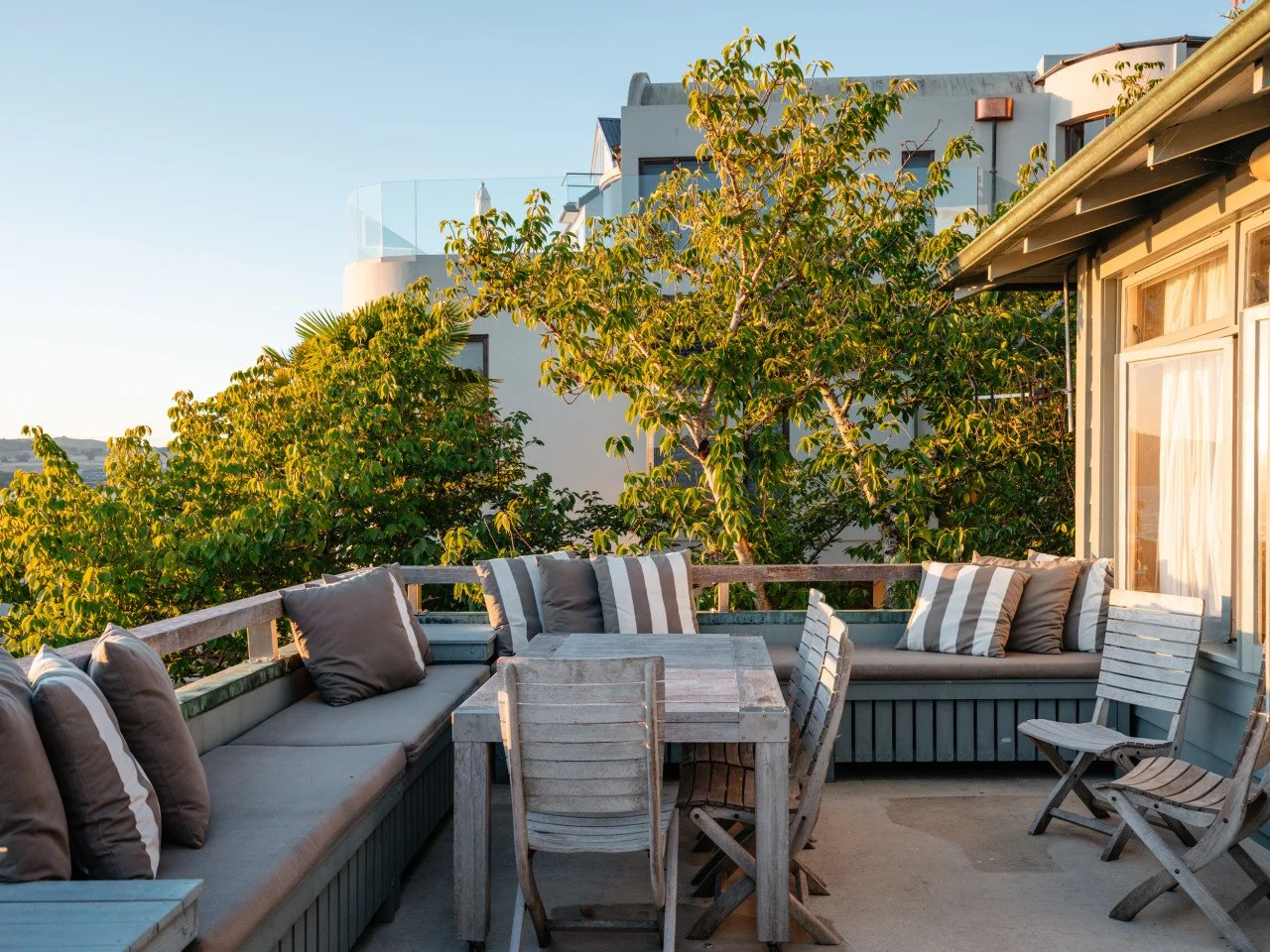 Outdoor balcony with gray wooden furniture including a table, chairs, and cushioned seats, surrounded by green trees and in front of a beige house with windows and curtains, under a clear sky.