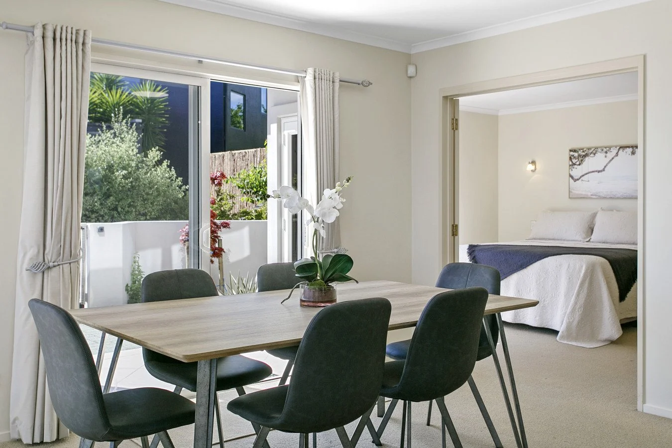 Dining area with a wooden table, six black chairs, and a white orchid centerpiece, with a view of a bedroom in the background.