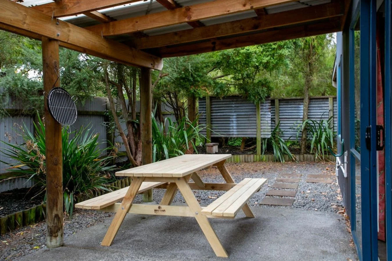Backyard with a wooden picnic table under a covered patio, surrounded by trees and plants, with a metal fence in the background and a gravel pathway.