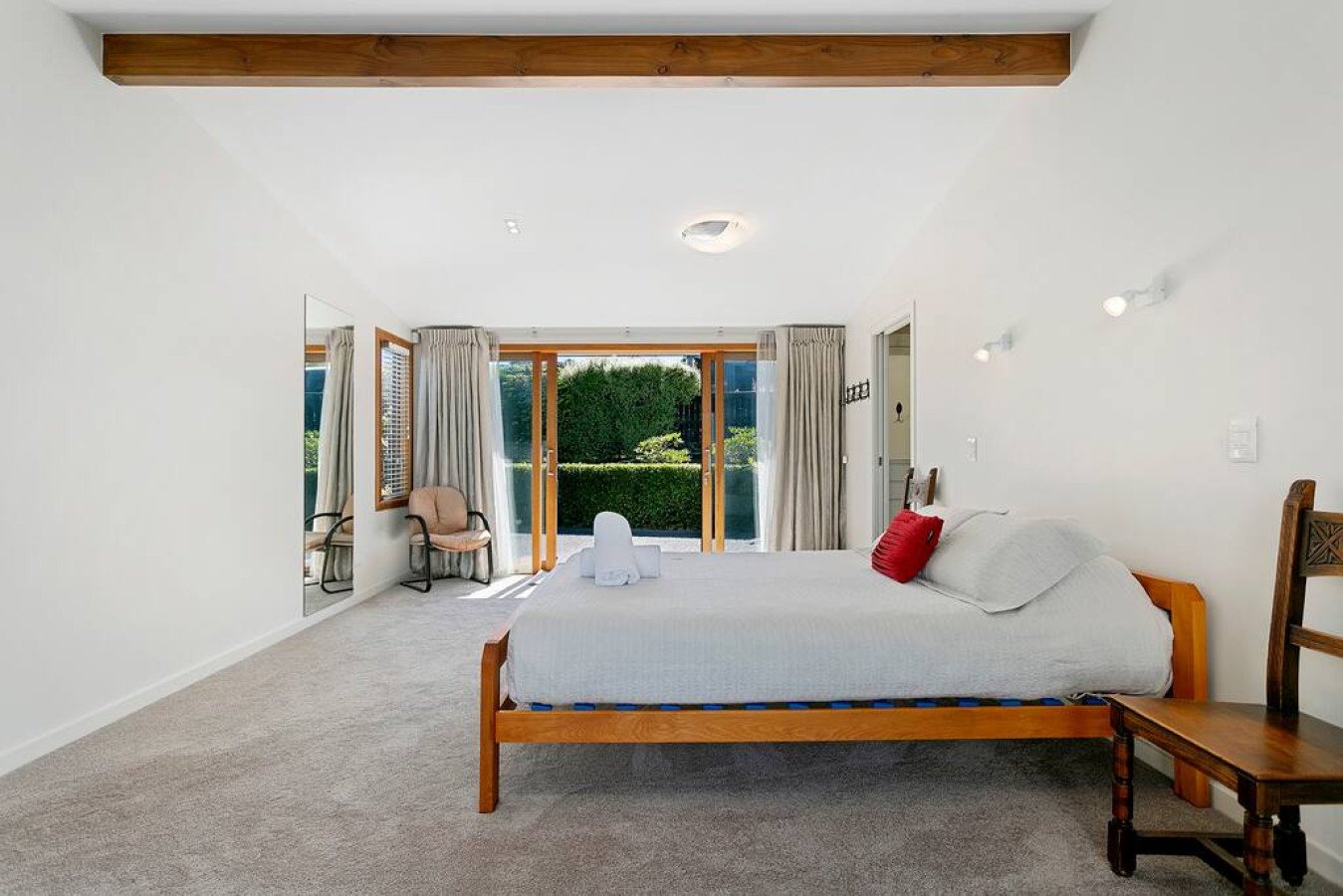 A bedroom with a large bed, a mirror, a small chair, and sliding glass doors leading to a garden.