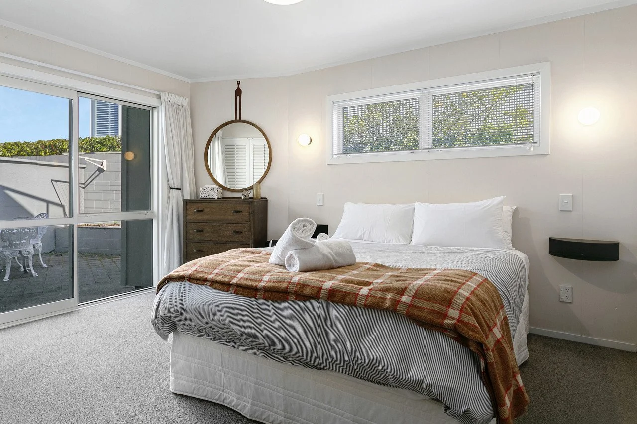 A bedroom with a large bed covered by a white comforter and a plaid blanket, two rolled towels on top, a chest of drawers with a mirror and decorative items, a sliding glass door leading to an outdoor patio, and a colorless window with blinds, with s